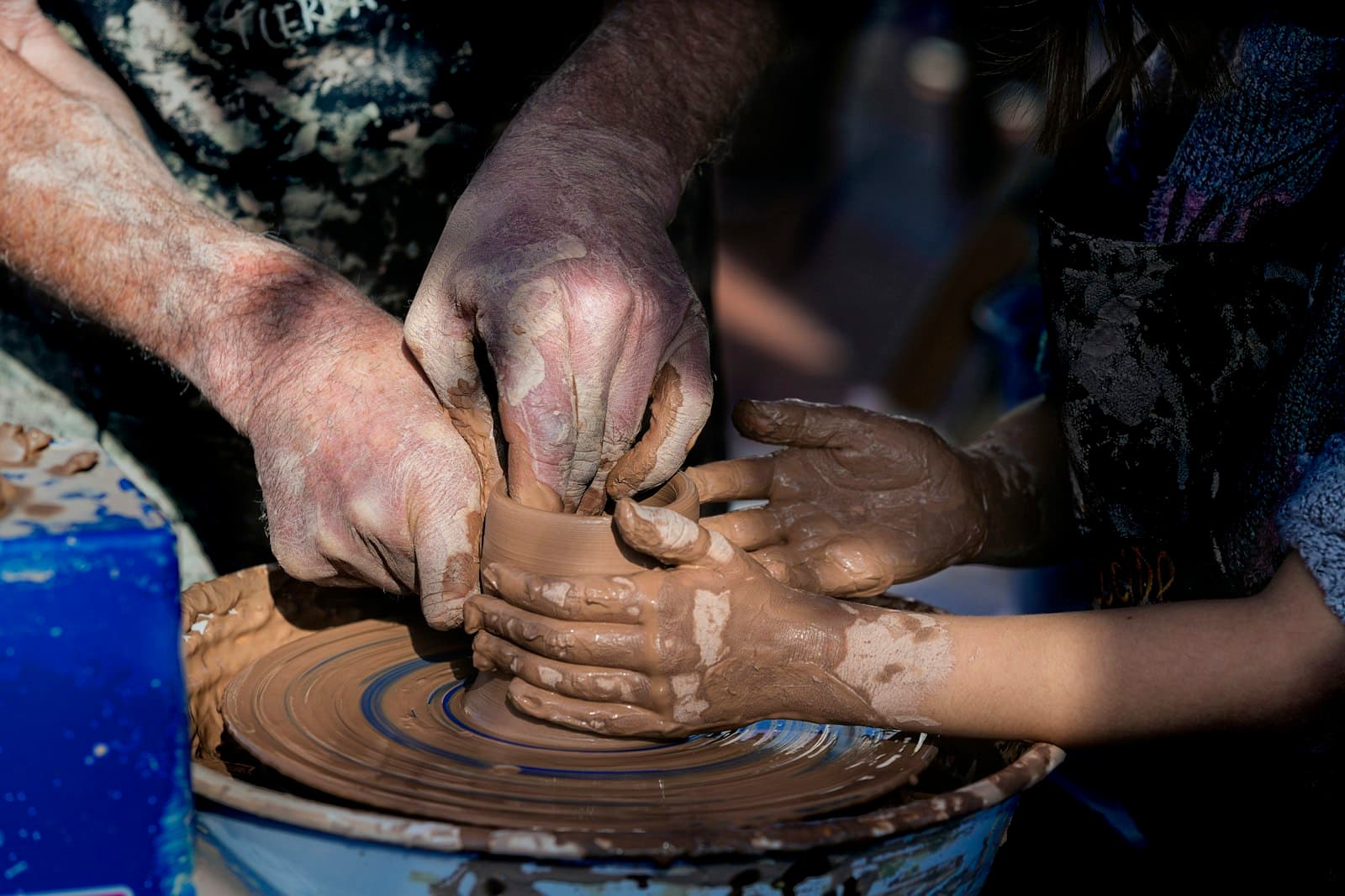 Two pairs of clay-covered hands shaping a pot together on a potter’s wheel